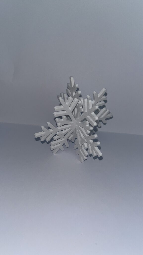 Weiße 3D-gedruckte Schneeflocke, aus zwei kreuzweise gesteckten Teilen, stehend vor hellem Hintergrund.