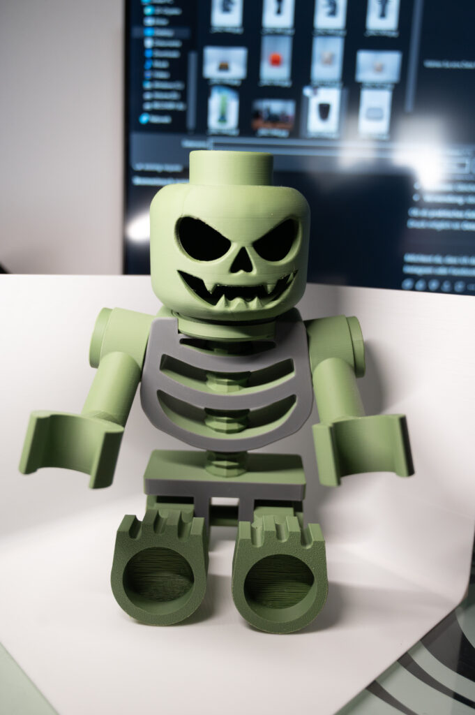 3D-gedruckte grüne Skelett-Minifigur mit bösem Grinsen, sitzend in einer weißen Fotobox vor einem Monitor.