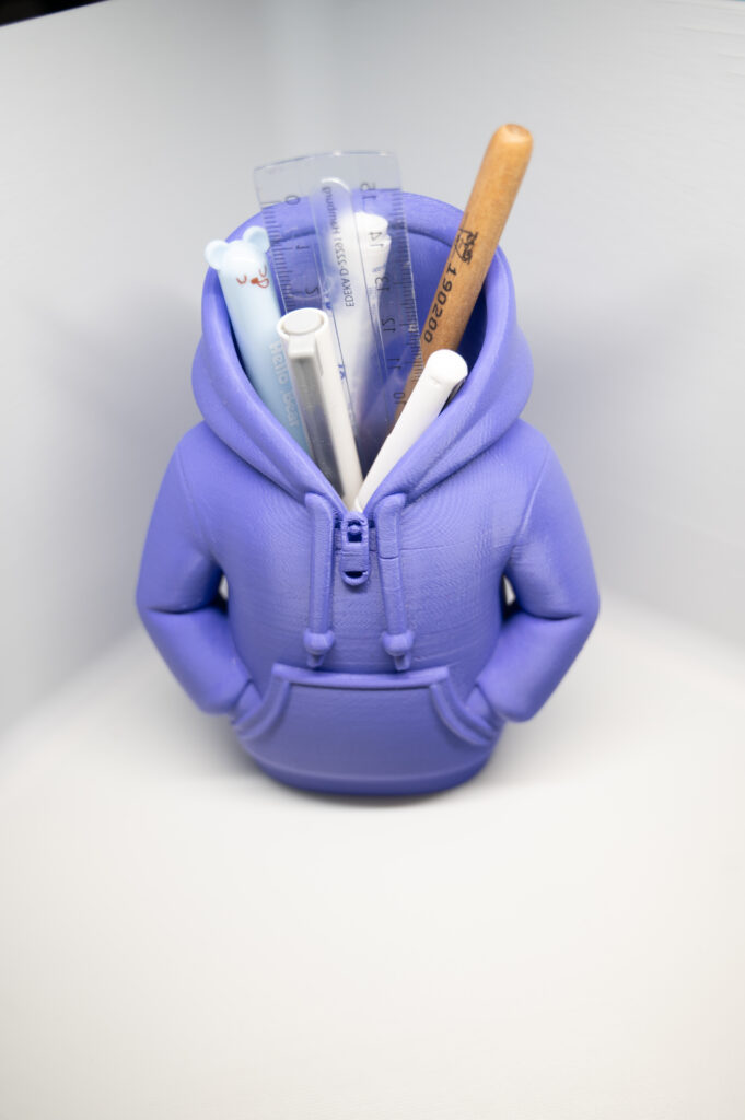 3D-gedruckter lilafarbener Hoodie-Stiftehalter mit Stiften, Lineal und Holzstift in einer weißen Fotobox.