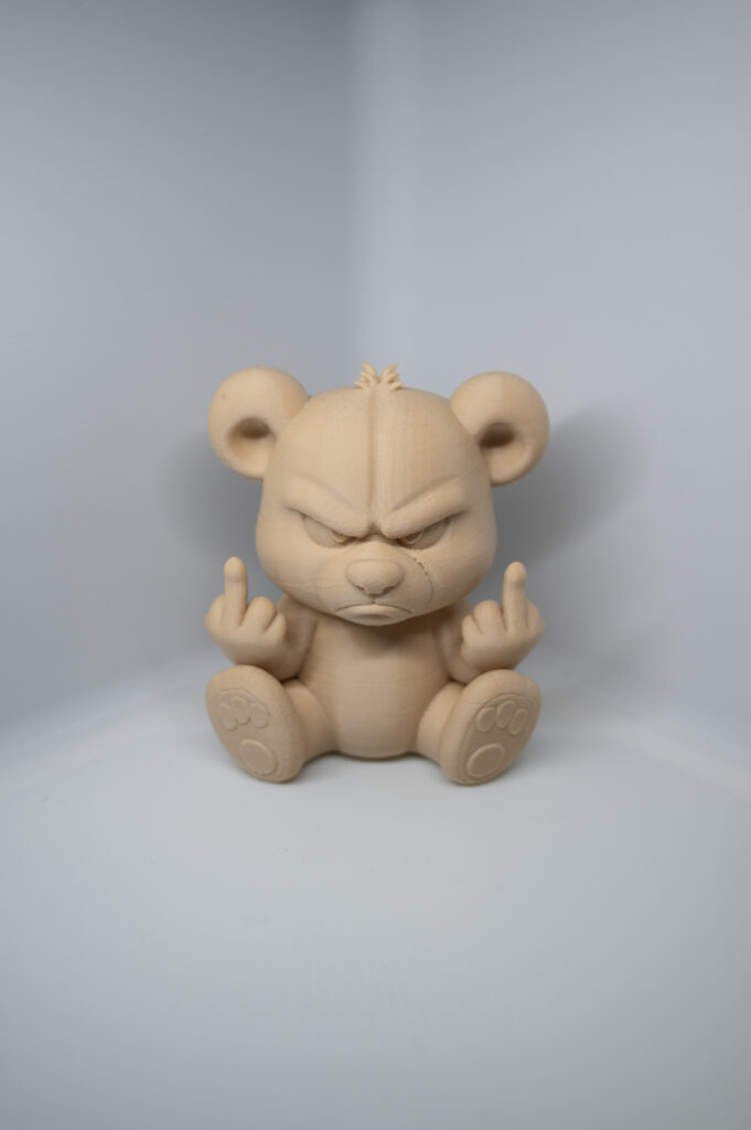 Beigefarbene 3D-gedruckte Bärenfigur mit grimmigem Gesichtsausdruck, die sitzend beide Mittelfinger zeigt, in einer weißen Fotobox.