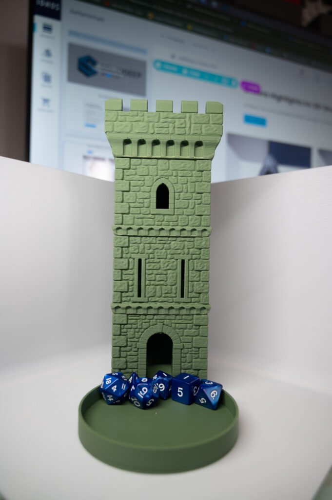 Grüner 3D-gedruckter Würfelturm im Burgdesign mit strukturierter Steinoberfläche und blauen Polyederwürfeln auf einer runden Schale in einer weißen Fotobox, dahinter ein unscharfer Monitor.