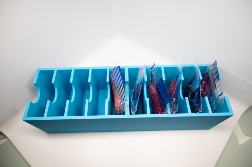 Blauer 3D-gedruckter Organizer mit mehreren Fächern, teilweise gefüllt mit stehenden, bunten Beutelchen in einer weißen Fotobox.